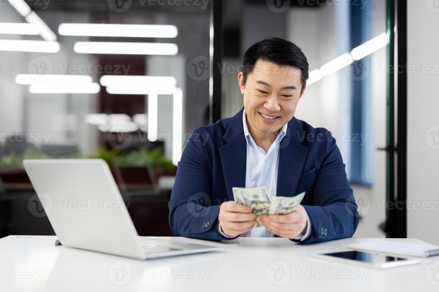 Smiling Asian businessman sitting at a modern office desk, counting US dollar bills. Concept of finance, success, and business lifestyle. photo