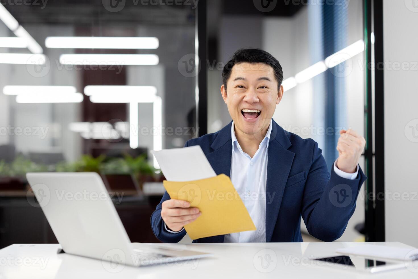 An Asian businessman in a blue suit sits at his desk in a modern office, ecstatically celebrating as he holds an envelope and reads its contents. photo