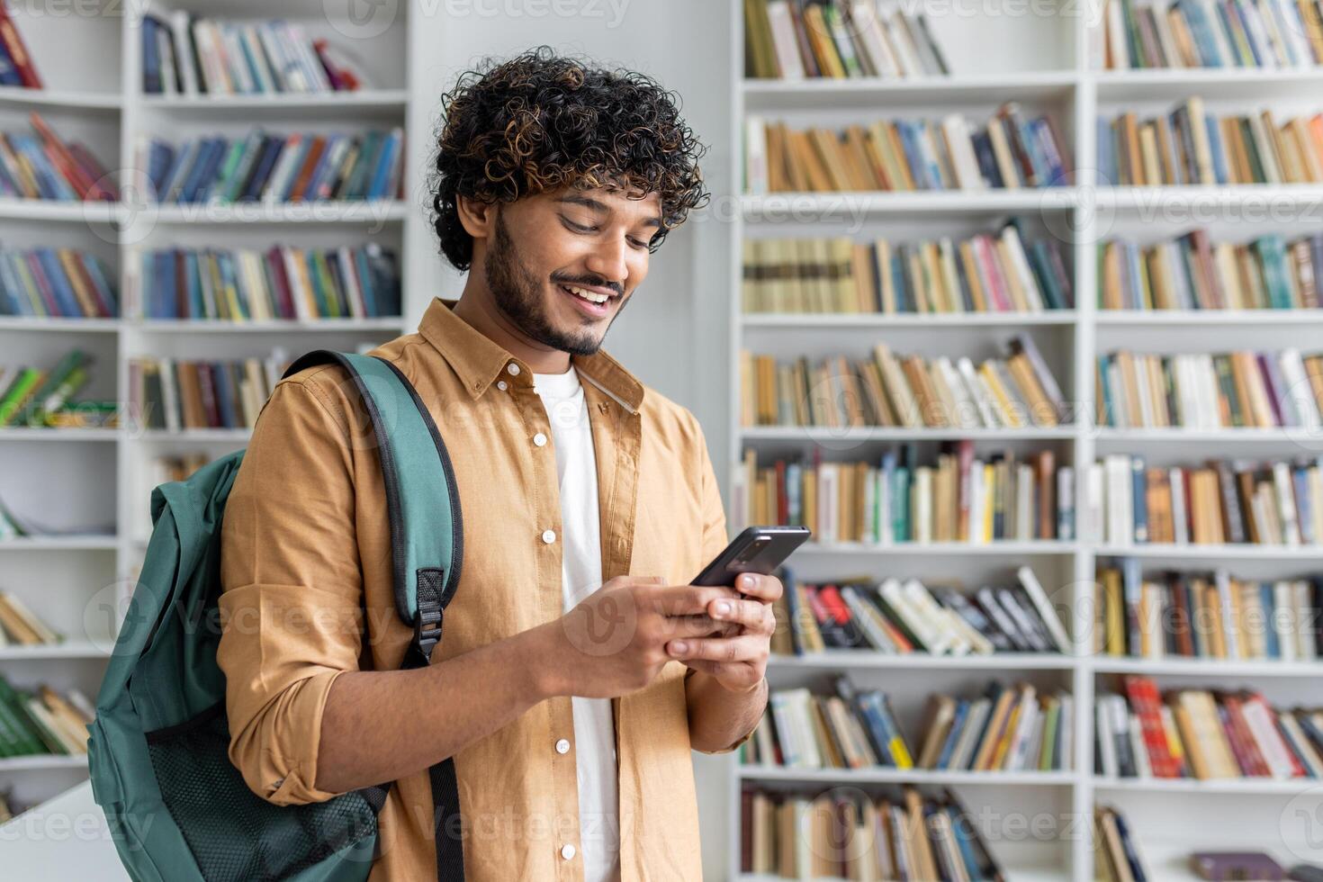 Positive middle eastern man wearing backpack slung over one shoulder standing in front of bookshelf and holding modern mobile phone. Happy male chatting with friends online in social medias. photo