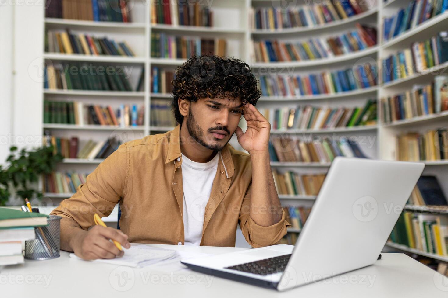 A focused young man is immersed in his studies at the library, surrounded by books, with a laptop for assistance. He seems to be preparing for exams, reflecting the life of a university or academy student. photo