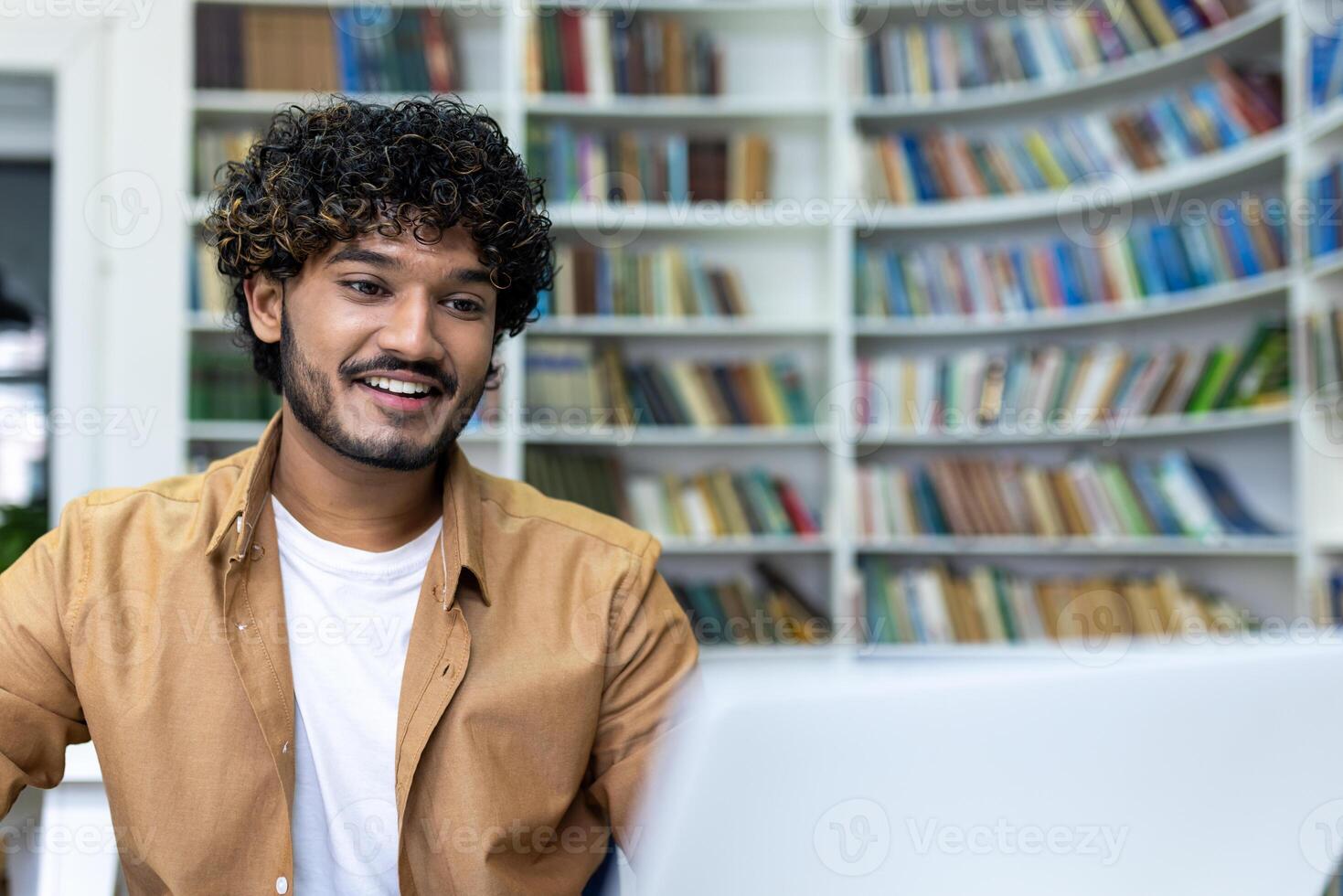 Young hispanic student studying inside university campus academic library among bookshelves, man smiling and using laptop. photo