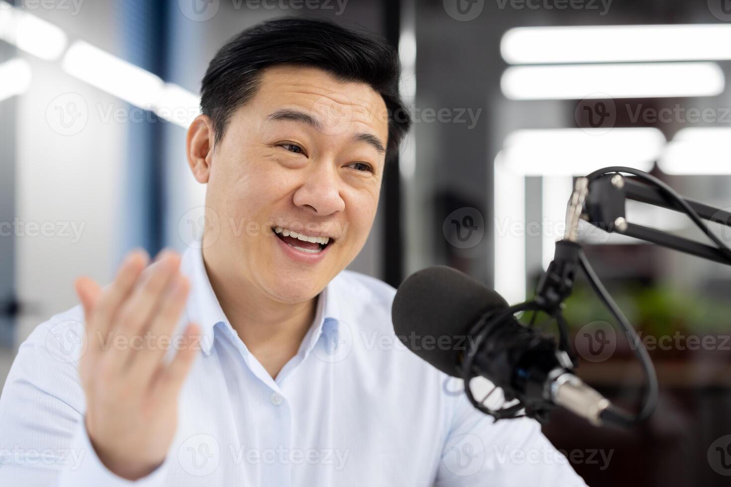 Close-up photo of a young Asian businessman in a white shirt standing in an office and smiling and talking into a microphone while gesturing ,with his hands.