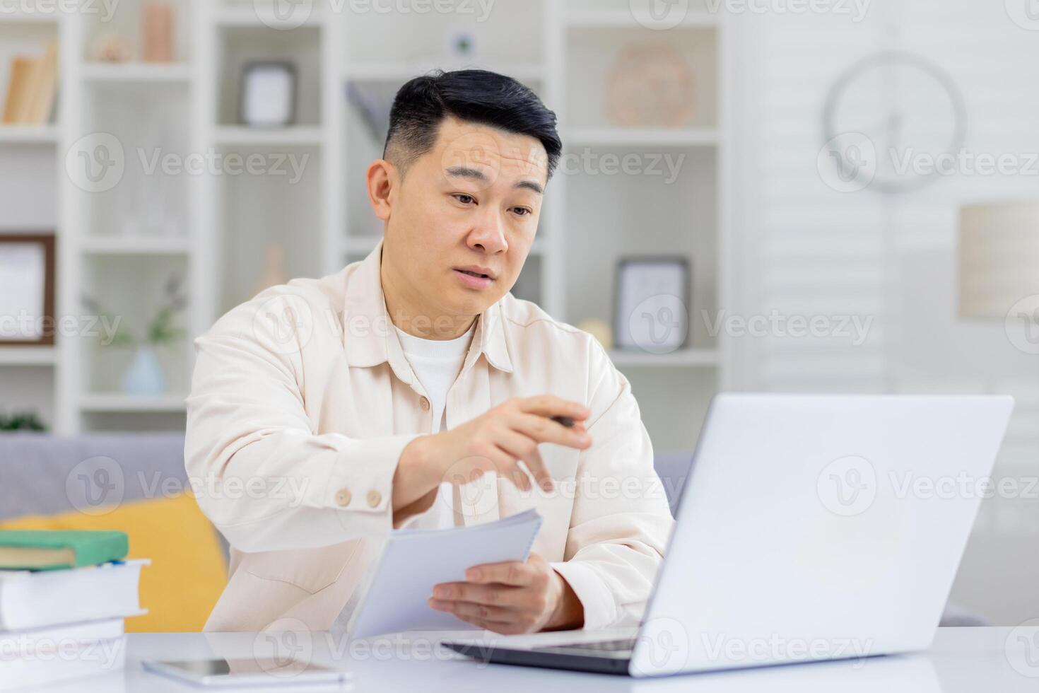 Asian young man sitting in the office at the table and talking on a call on the laptop, holding a notebook and a pen in his hands. photo