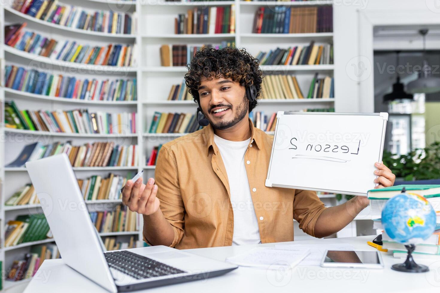 A cheerful male tutor wearing a headset teaches an online class, holding a whiteboard with 'lesson' written backwards, symbolizing remote education challenges and solutions in a home office setup. photo