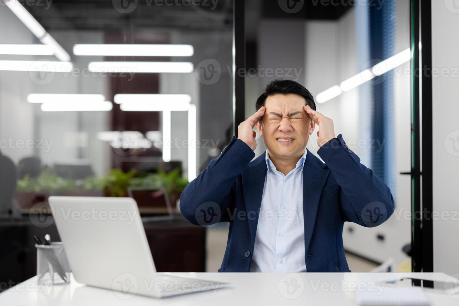 A man has a headache at the workplace inside the office, an Asian businessman in a business suit is sick, has a severe migraine, the boss is sitting at a desk with a laptop. photo
