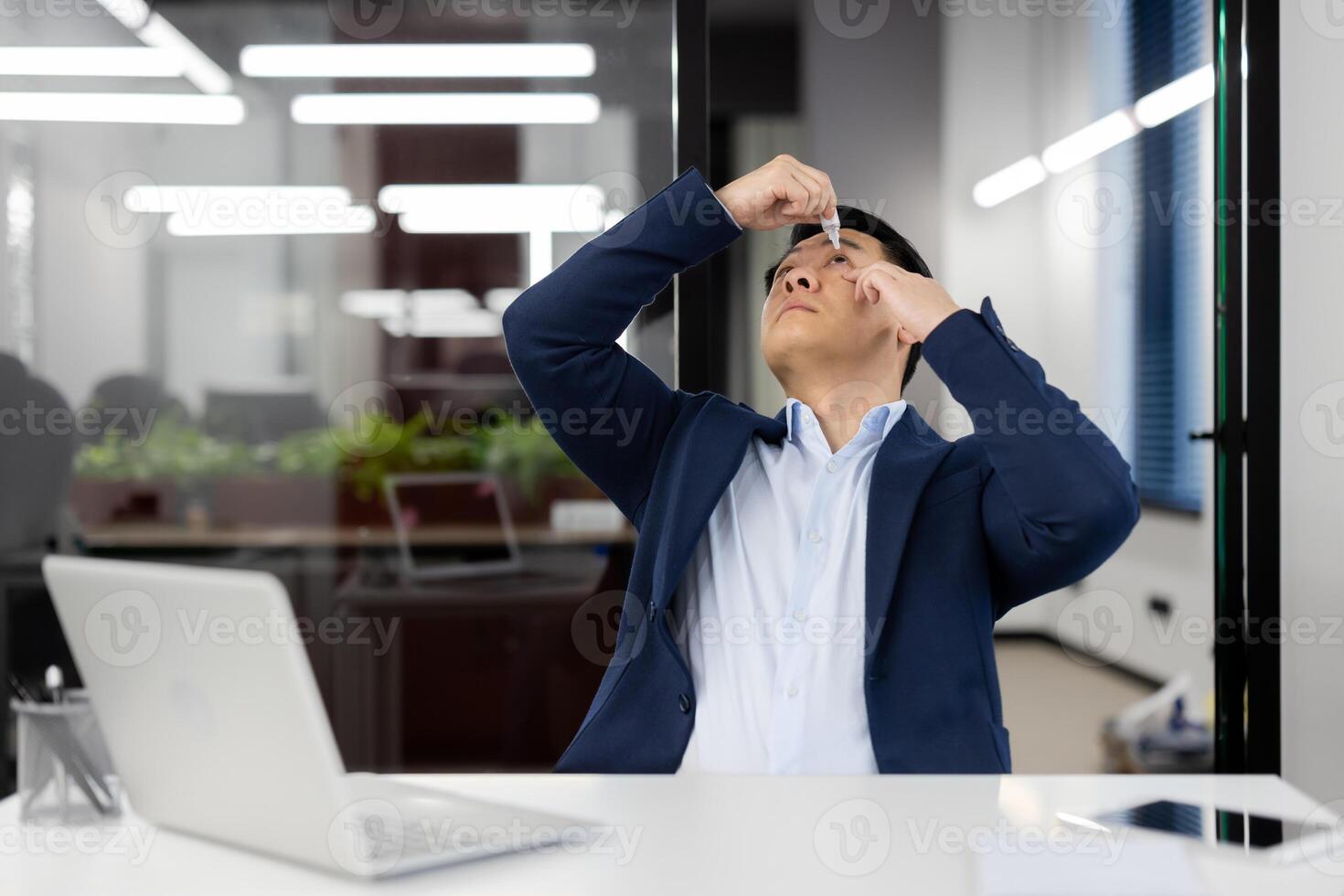 Asian young businessman instills his eyes with medicine drops for inflammation and overfatigue. He sits at his desk in the office in a suit and works overtime. photo