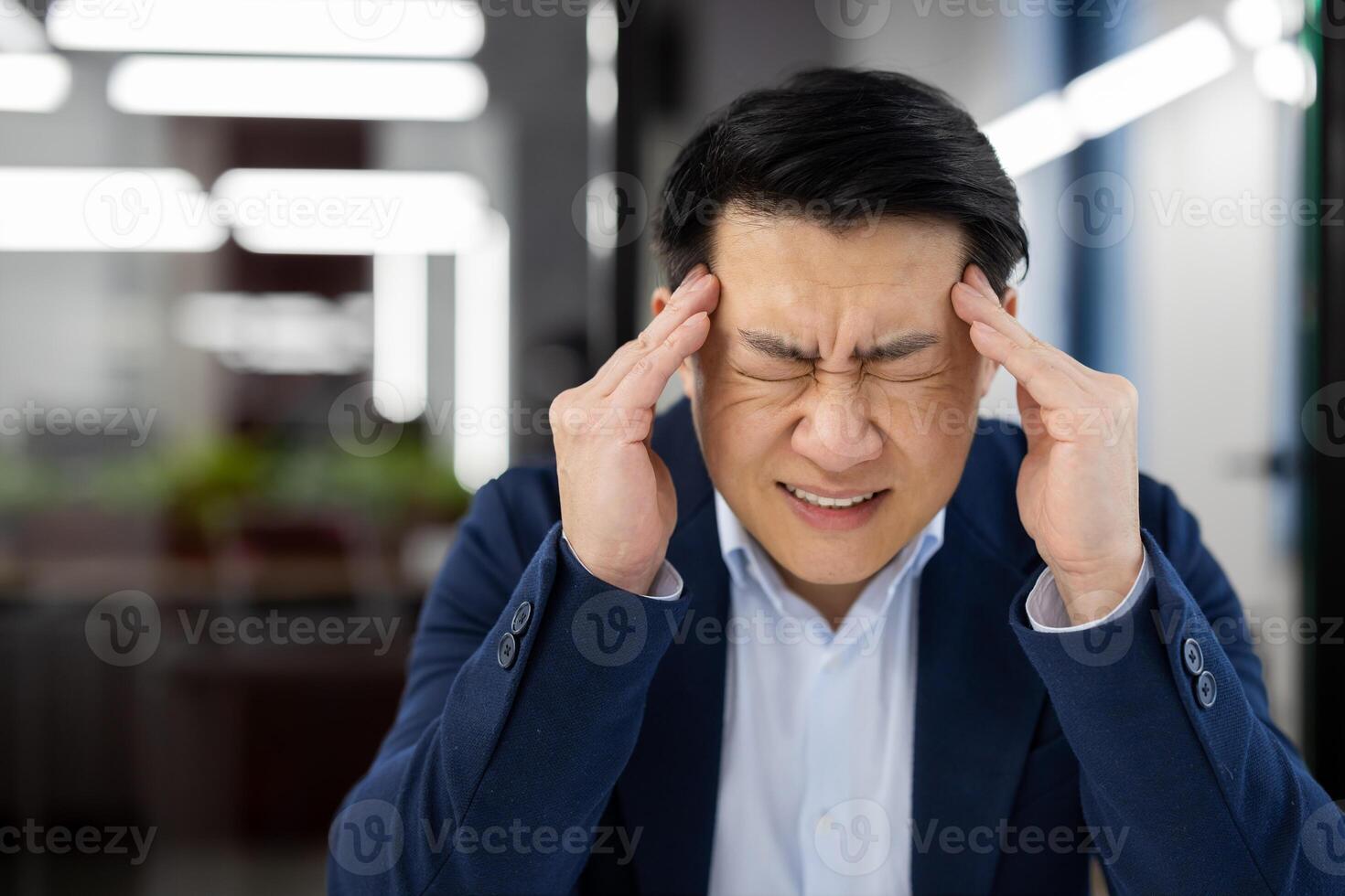 A man has a headache at the workplace inside the office, an Asian businessman in a business suit is sick, has a severe migraine, the boss is sitting at a desk with a laptop. photo