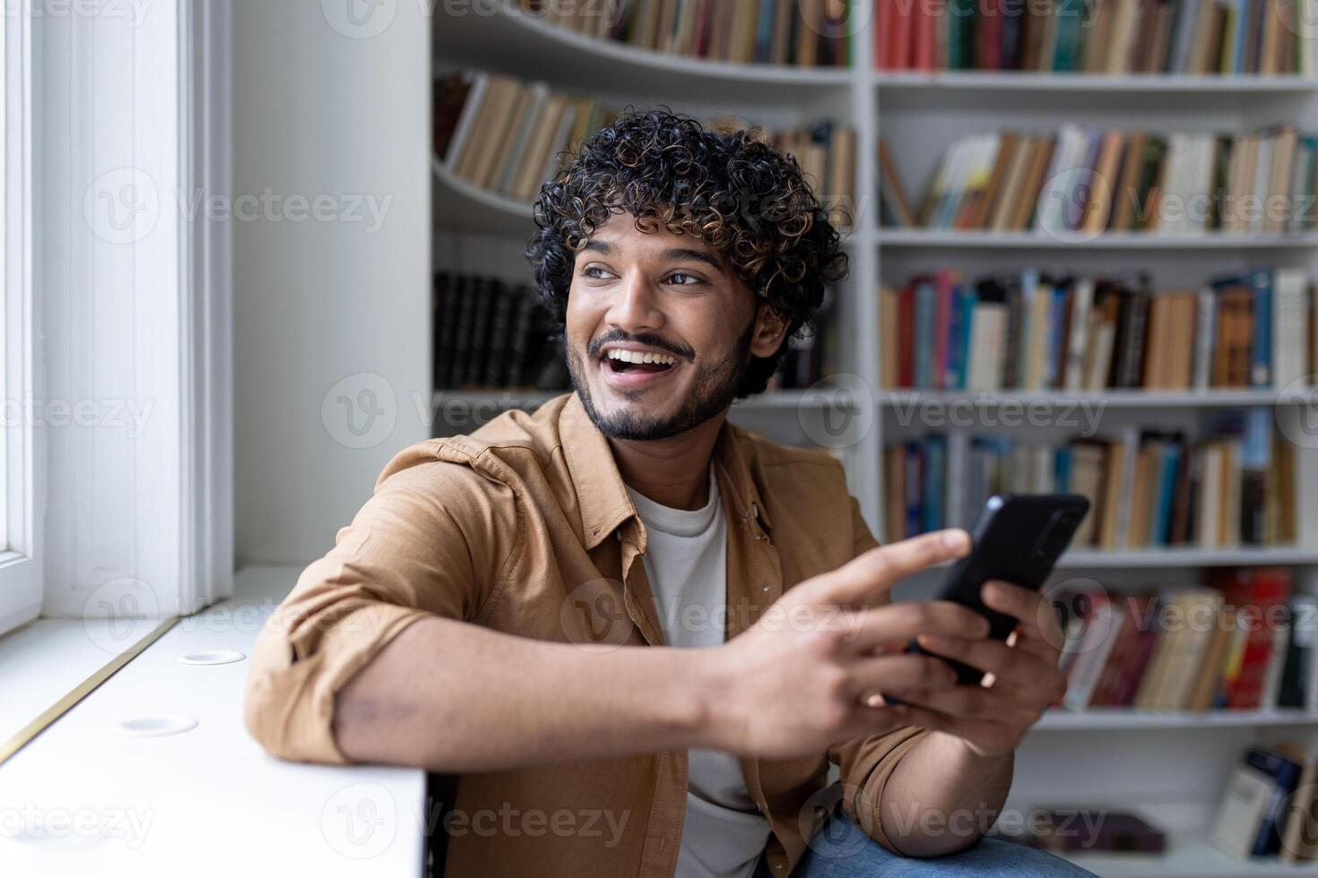 Young student among bookshelves studying and smiling, hispanic man holding phone, using online application. photo