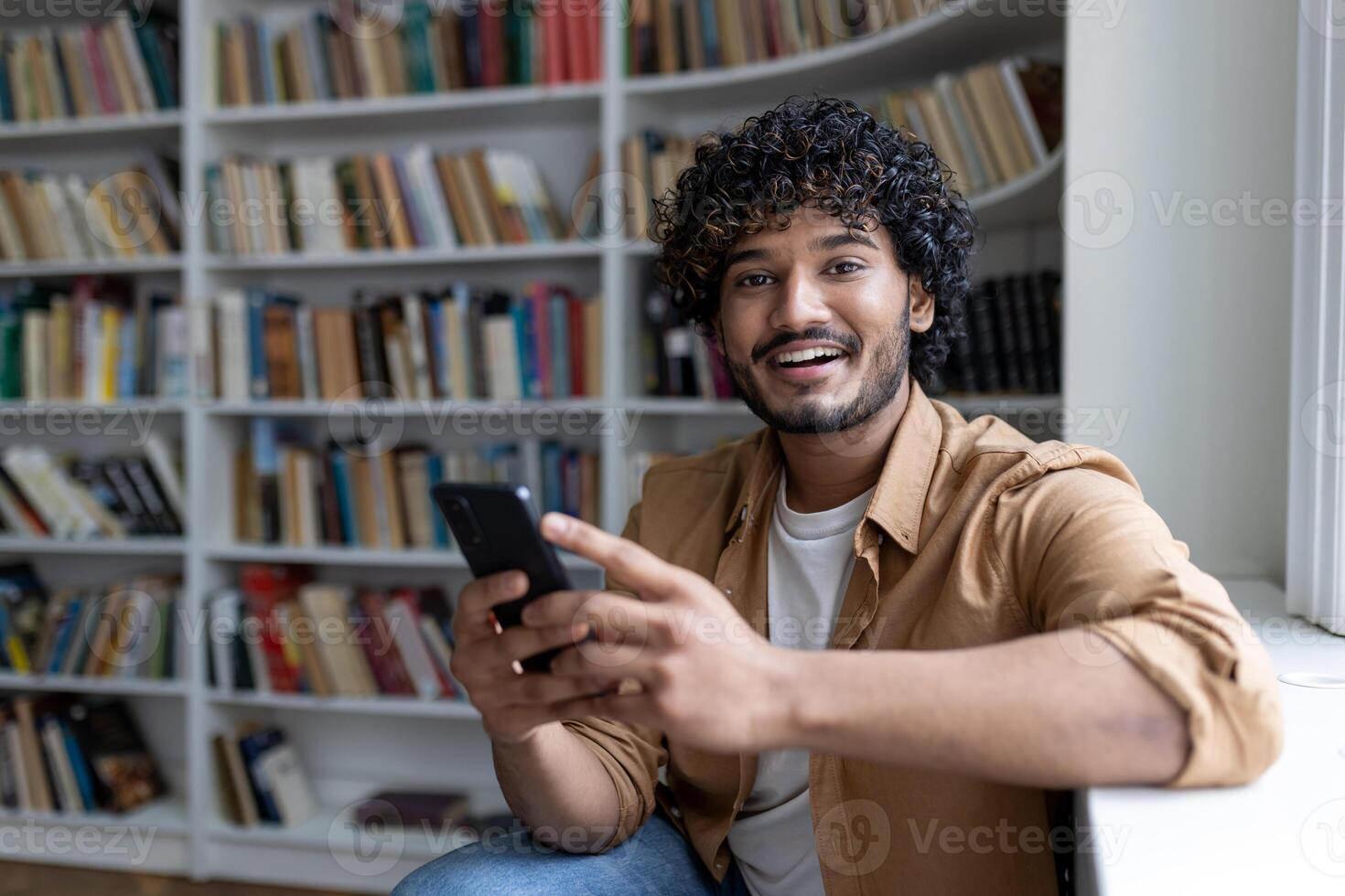 Portrait of young hispanic student, man holding phone smiling and looking at camera, studying inside public library, on university campus. photo