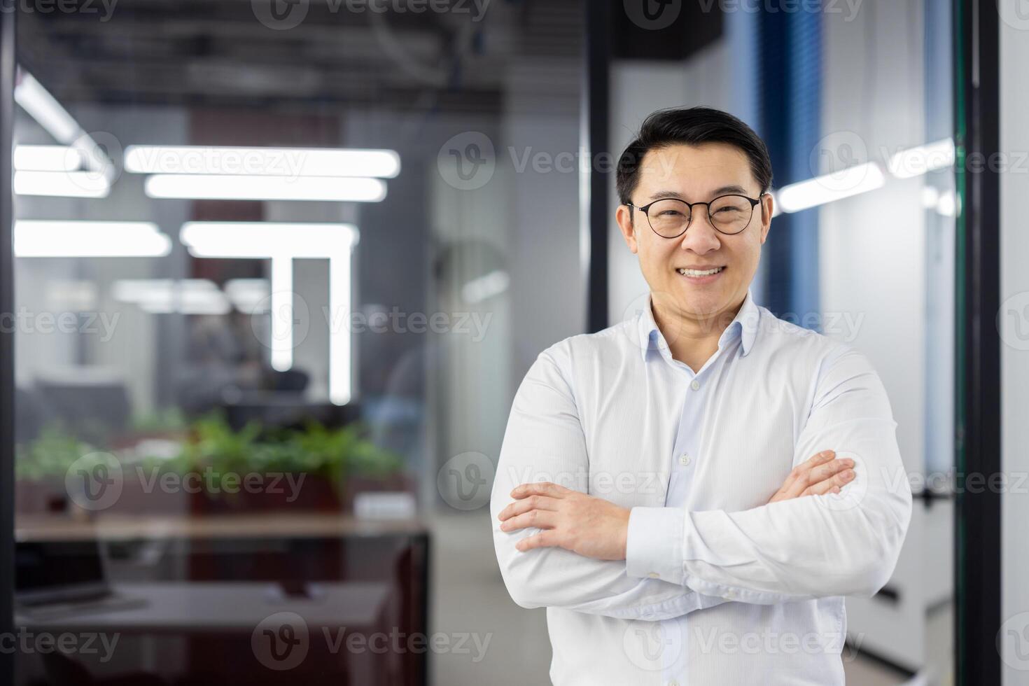 A professional Asian businessman stands confidently with arms crossed, smiling in a modern office setting. photo