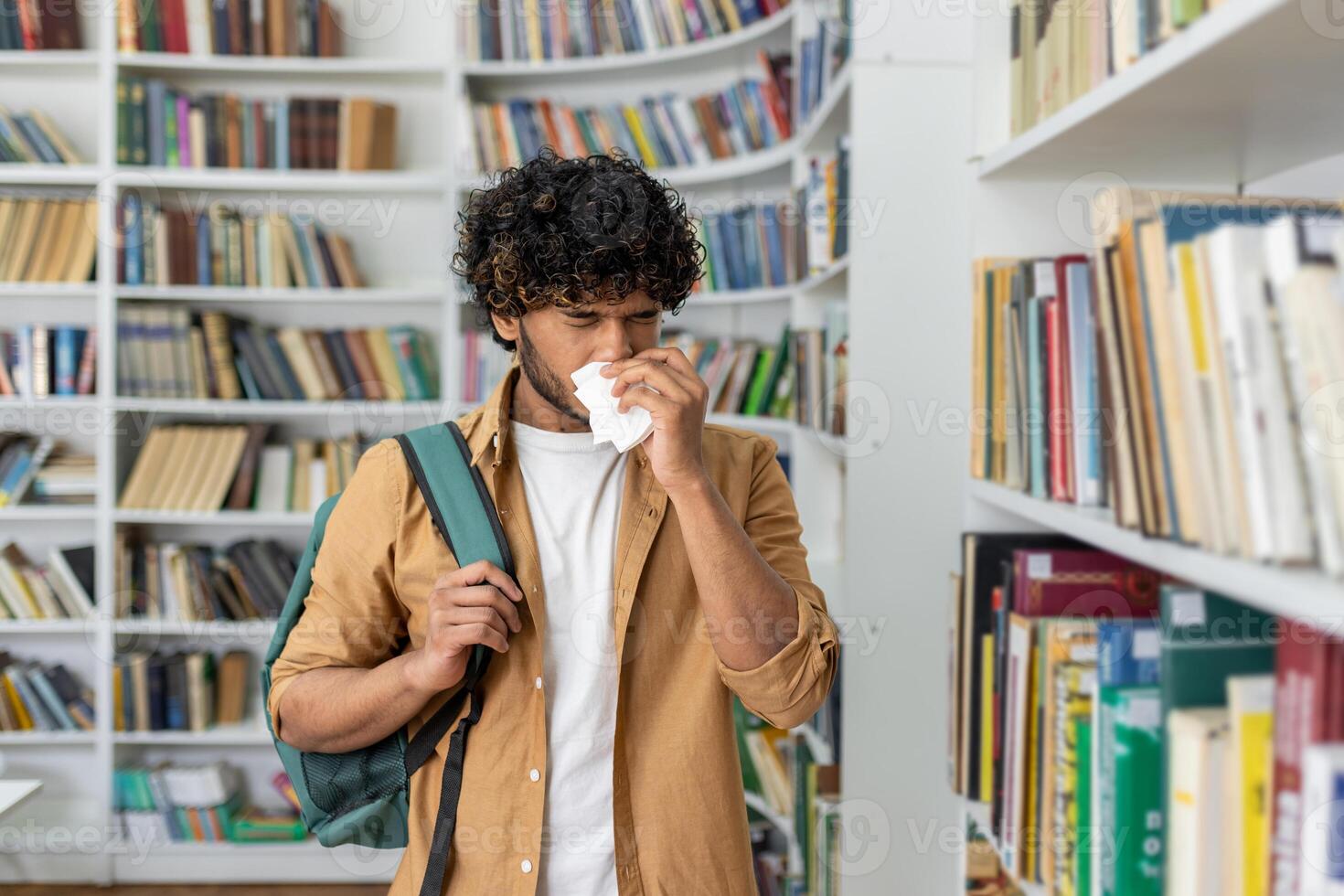 Unhealthy man in casualwear using paper napkin while sneezing and standing by aisle with collection of novels. Sick islamic male struggling with dust allergy in book shop while holding shoulder bag. photo
