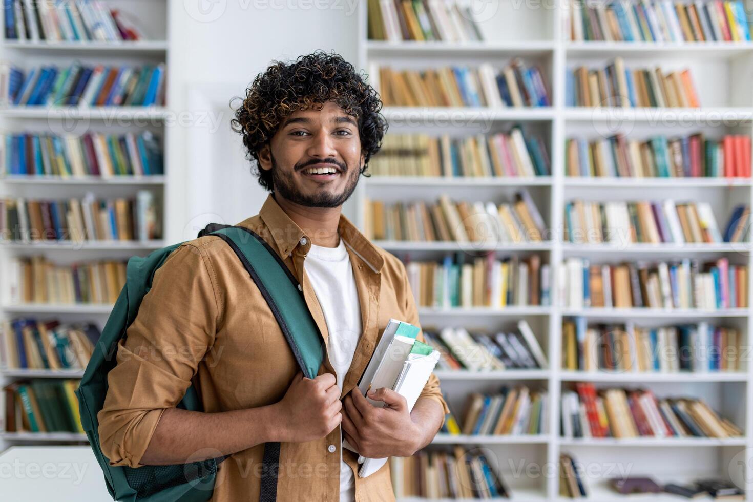 Portrait of happy satisfied african american student with curly hair, guy with books and backpack standing in university academic library smiling and looking at camera. photo