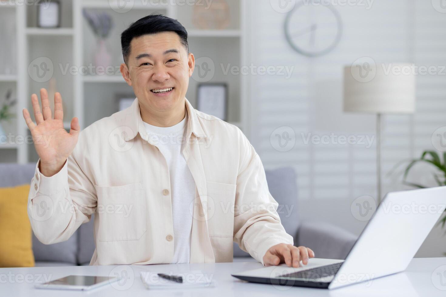 Portrait of a young smiling Asian man working in the office using a laptop, sitting at the table, waving and greeting the camera. photo