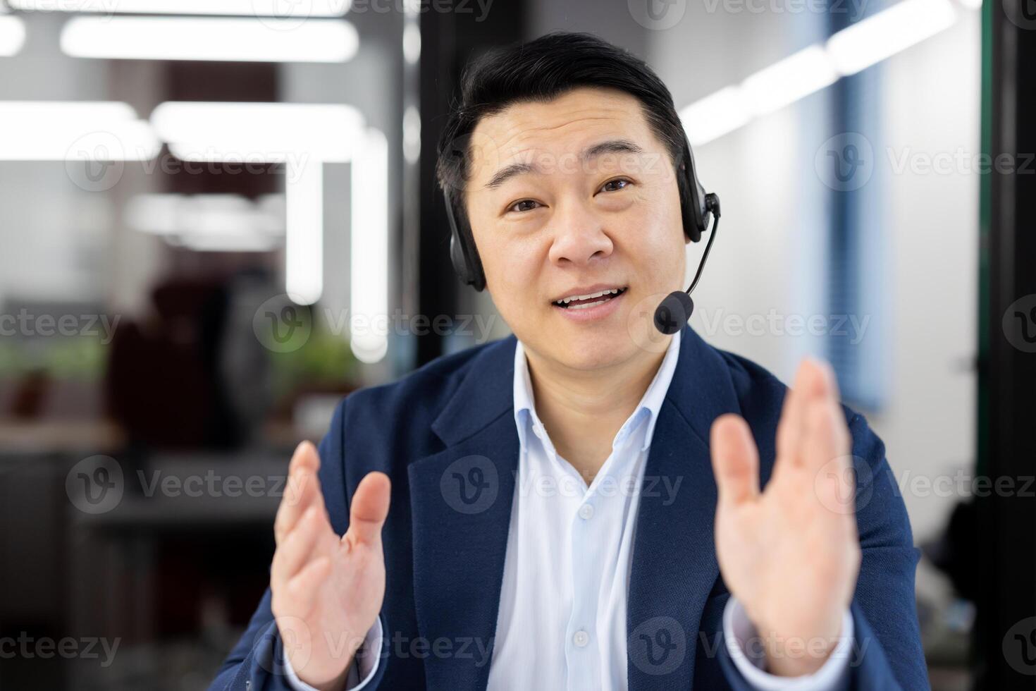 Engaged Asian businessman in a suit using a headset for a conference call in a modern workspace expresses positivity. photo