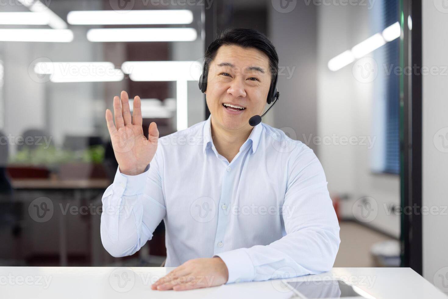 Friendly korean entrepreneur in headset with mic and shirt raising hand for greeting gesture while sitting by desktop. Positive office worker using computer for online virtual business communication. photo