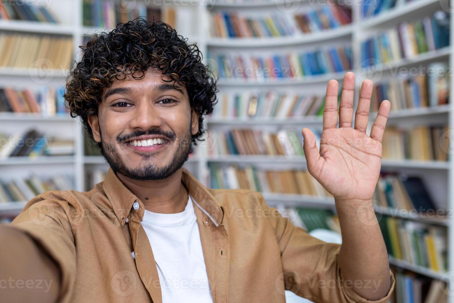 Young hispanic male student chatting online via call in the library. Smiles at the camera, shoots a blog, waves, studies. Close-up photo. photo