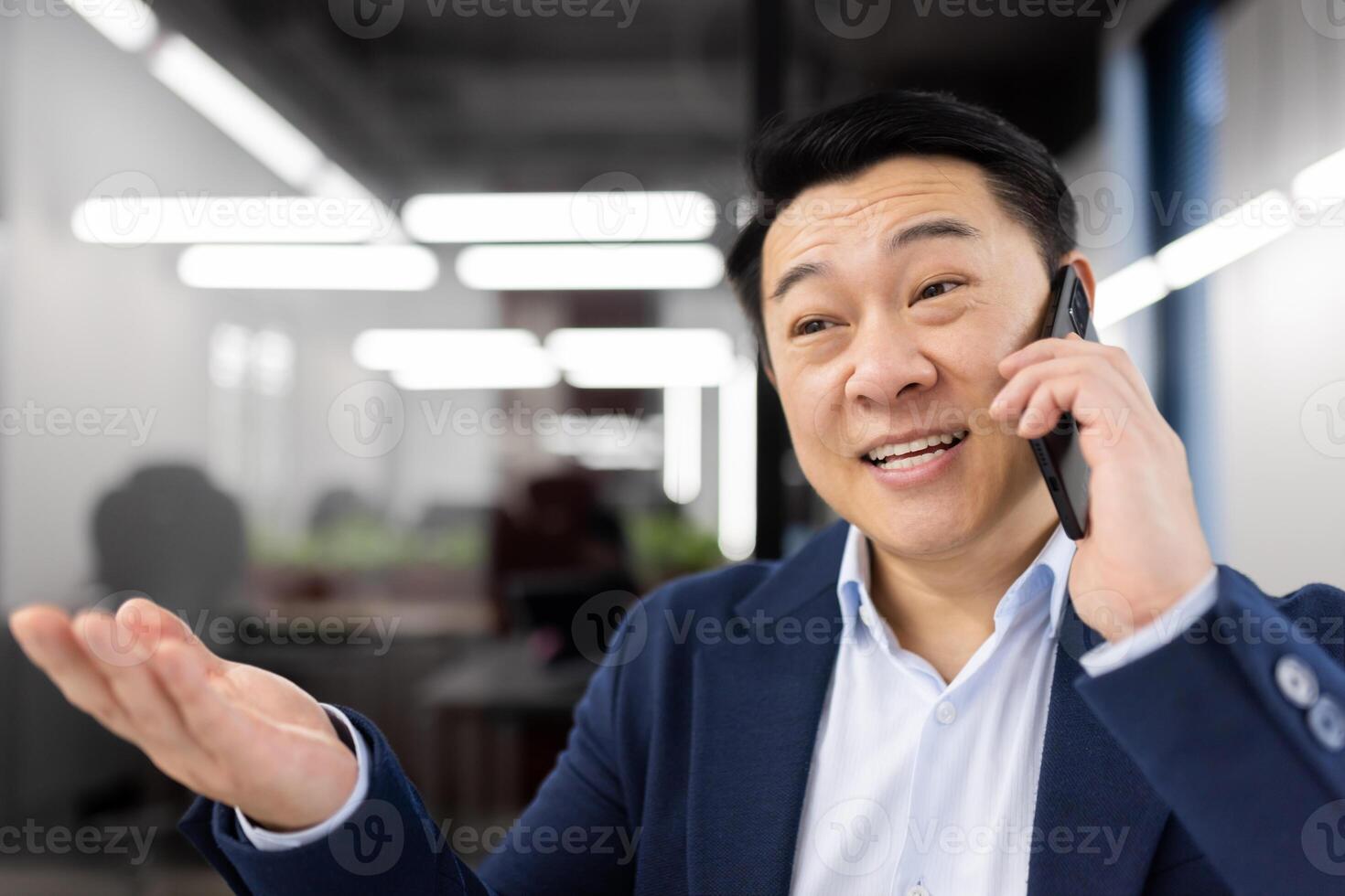 Cheerful Asian businessman engaged in a phone conversation in a modern office setting. He appears happy and enthusiastic, gesturing with his hand. photo