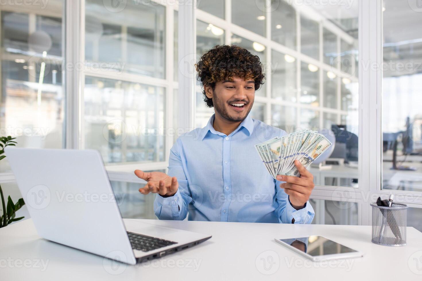 Cheerful young man in a light blue shirt joyfully holding and counting a stack of money at his office desk, with laptop and tablet present. photo