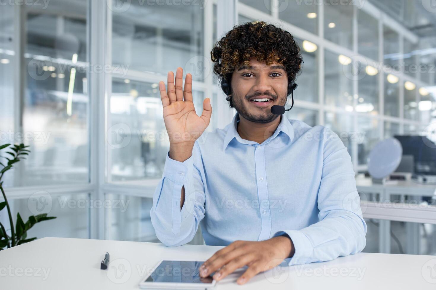 Hindu male wearing headset with microphone and raising hand in greeting gesture while sitting by desk with tablet. Optimistic support operator welcoming new colleague in sharing working space. photo