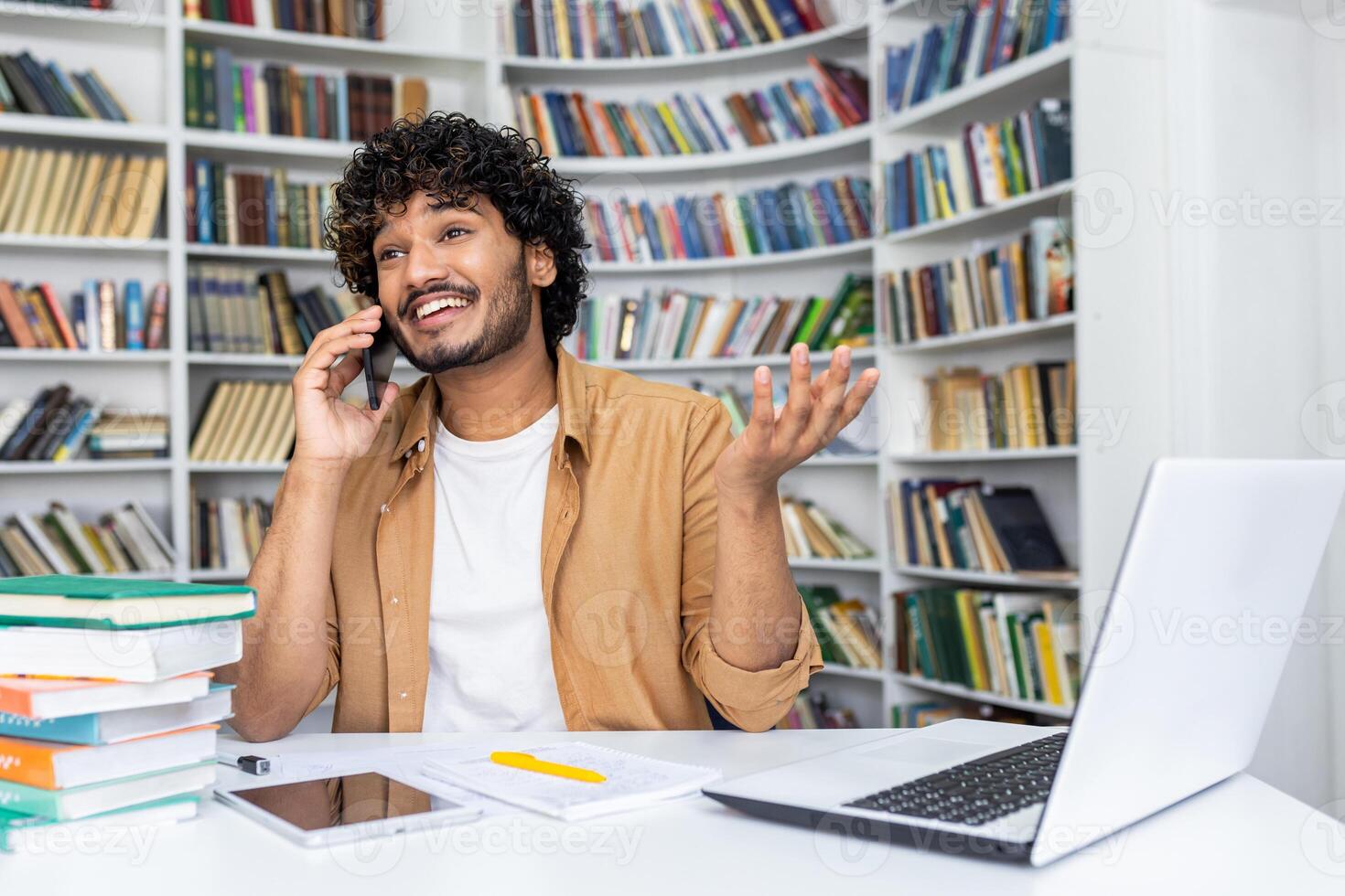 Happy young man with curly hair makes a phone call while working on his laptop in a home office with bookshelves in the background. photo