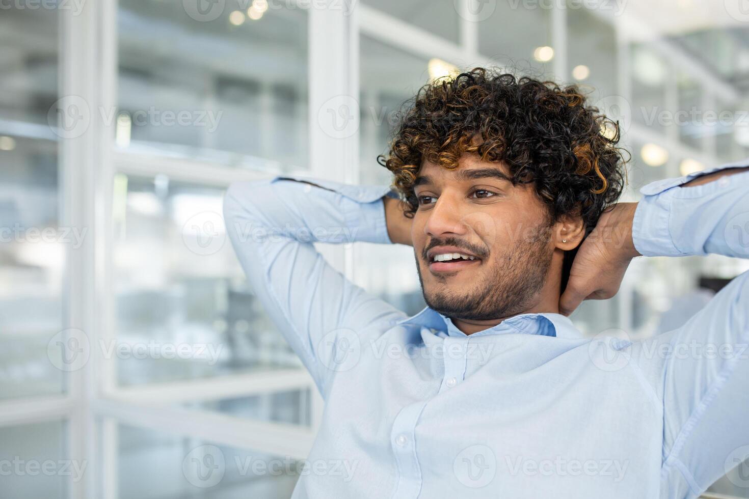 Portrait of young unshaved man in formalwear holding hands behind head while leaning on back of chair. Positive worker looking forward and daydreaming about upcoming vacation on blurred background. photo