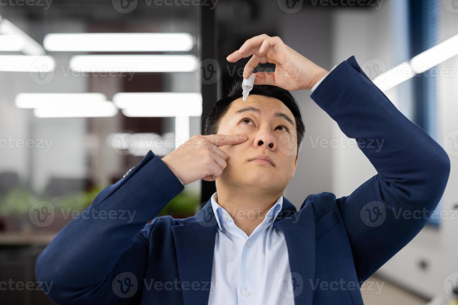 Close-up photo of a young Asian businessman who drips drops of medicine into his eye from overwork and illness. Located in the office behind the workplace.