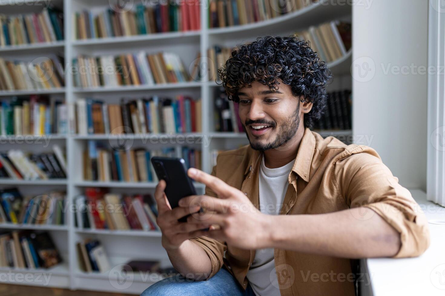 Young student among bookshelves studying and smiling, hispanic man holding phone, using online application. photo
