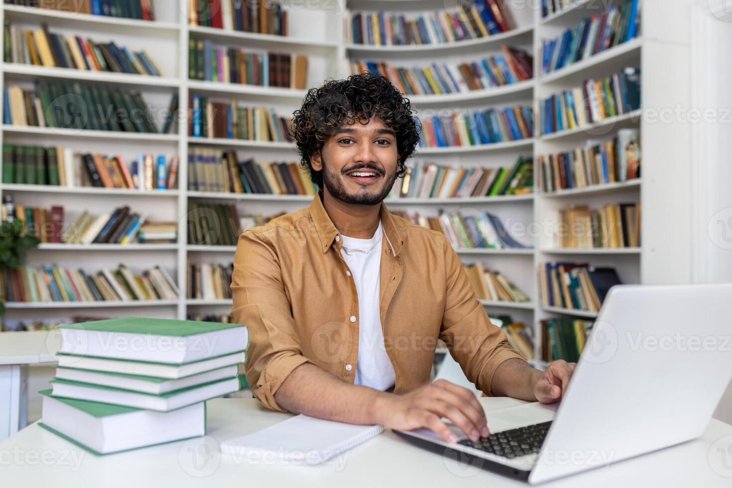Portrait of a smiling young Indian man studying and working in a library at a laptop. Sitting at a table with books and looking at the camera. photo