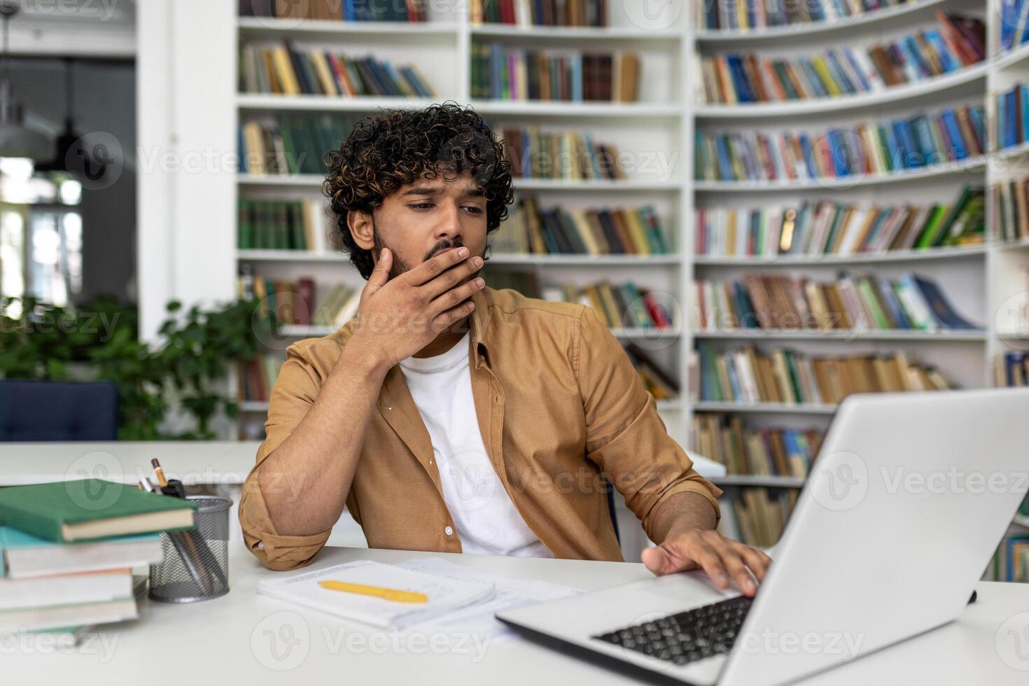 A concentrated male student sits in a library, hand on chin, deep in thought while studying on his laptop for upcoming exams. Books surround him, symbolizing academic research and learning. photo