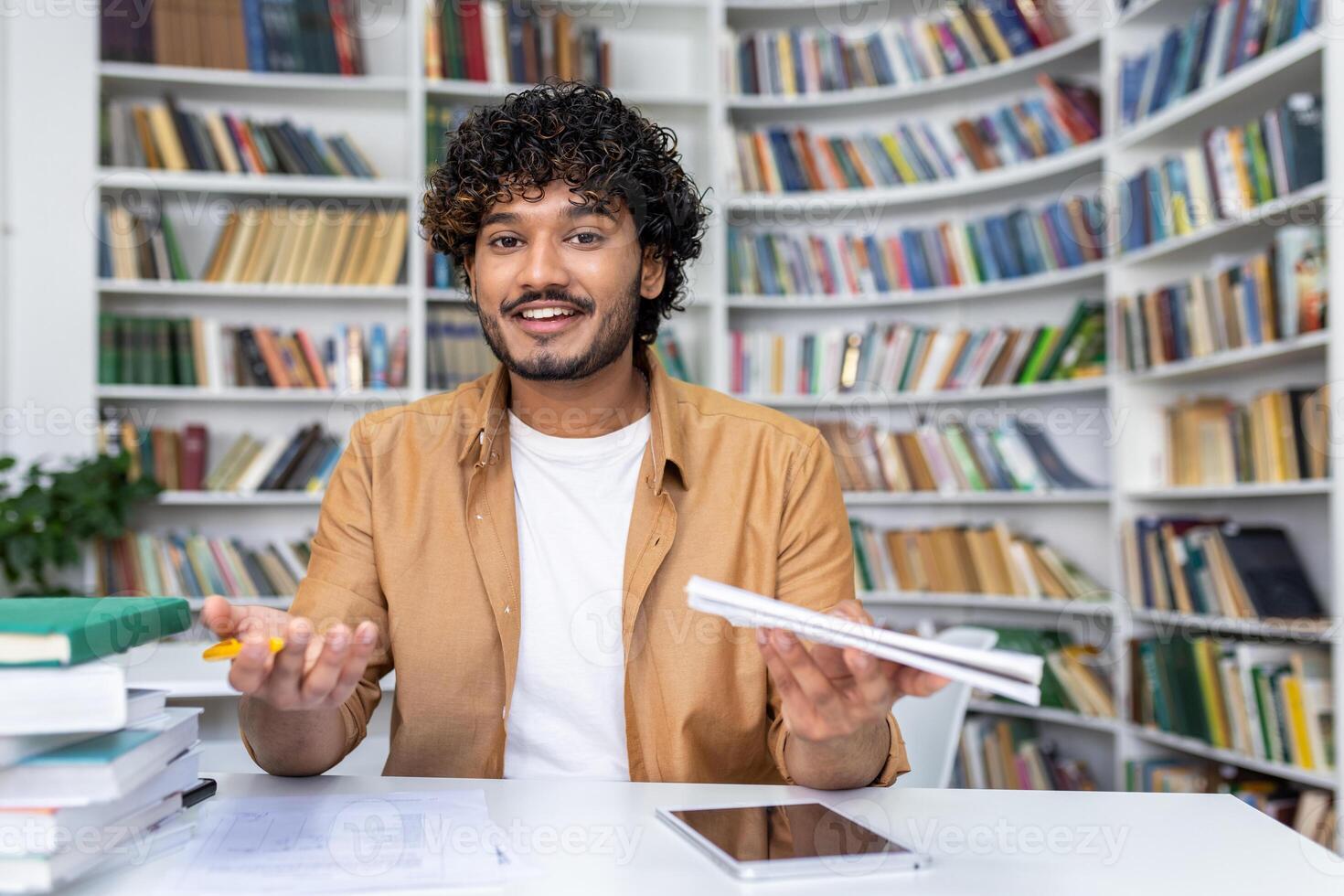 Young adult, confused and overwhelmed, sits at a desk cluttered with books and papers in a library setting. photo
