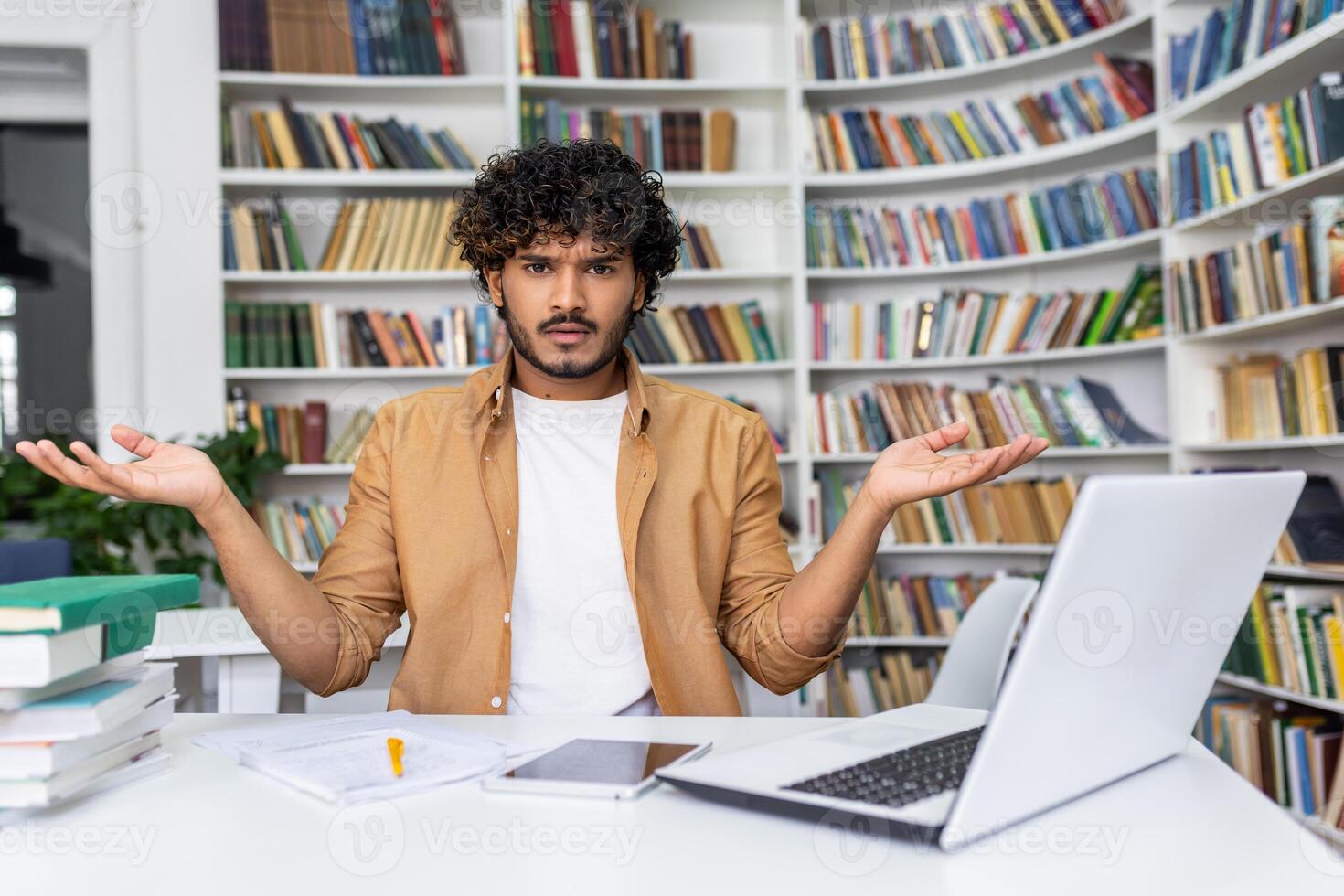 Surprised muslim man sitting at desk with modern laptop and papers surrounded by bookshelves. Male wearing white shirt and light brown jacket gesturing with both hands raised and palms up indoors. photo
