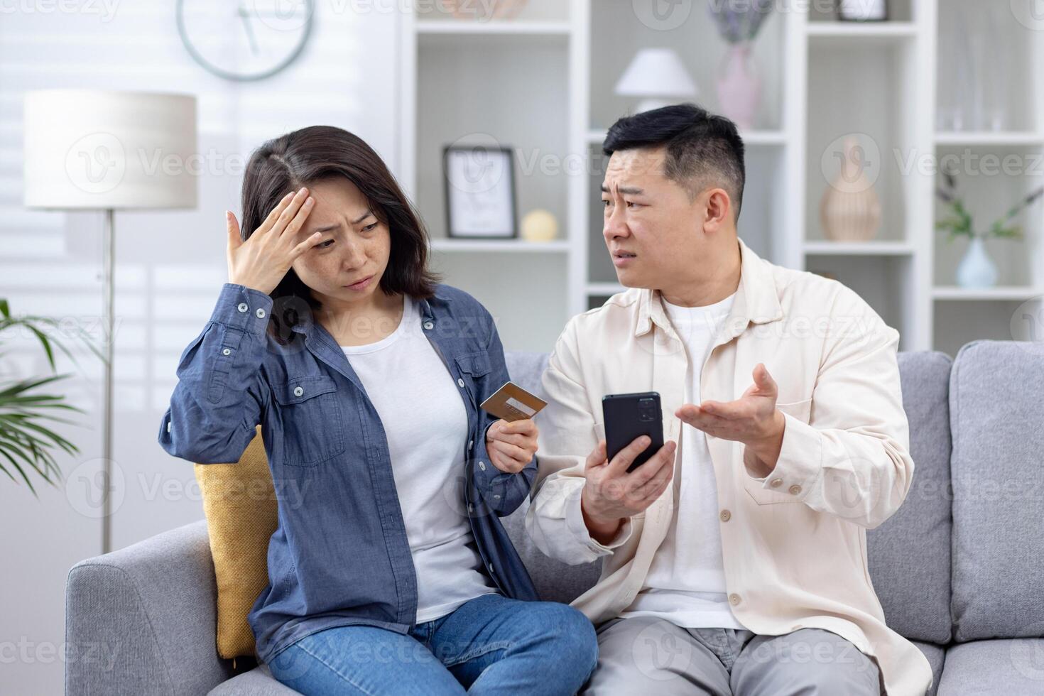 Asian couple sitting on couch discussing credit card fraud while using smartphone, concerned expressions photo