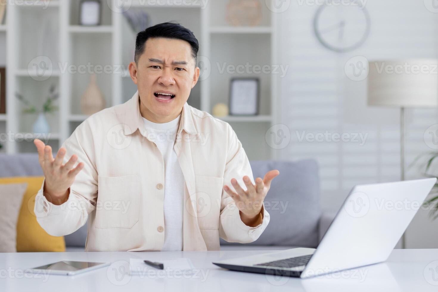 Man expressing frustration during a call meeting at home. He is seated at a desk with a laptop and paperwork. photo
