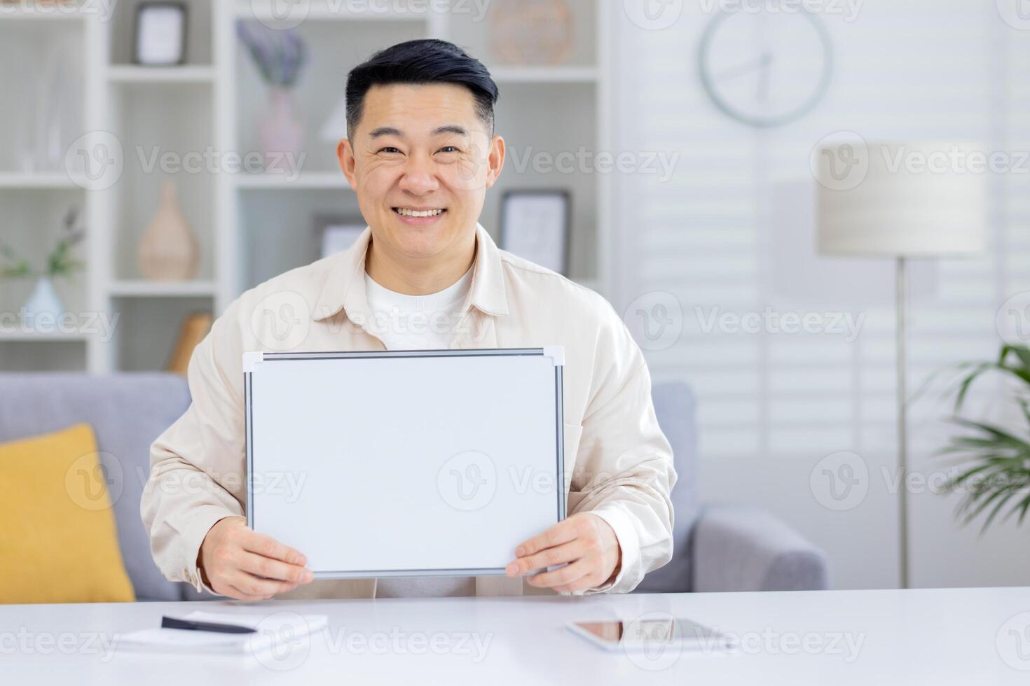 Portrait of a smiling Asian man sitting at a desk in a modern light office and holding a white blank clipboard in his hands, pointing and looking at the camera. photo