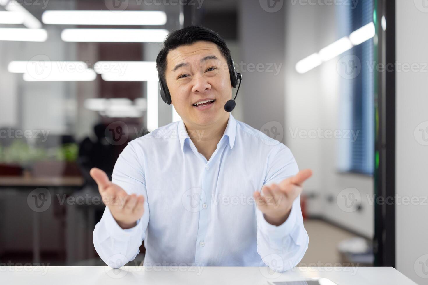 Webcam view, Asian support worker in shirt with headset smiling and looking at camera, businessman working inside office, consulting clients remotely, remote meeting with colleagues partners. photo