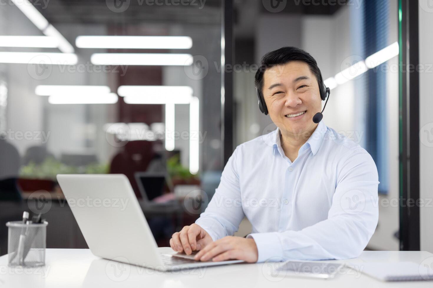 Positive office clerk in white shirt and black headset sitting by desk with portable computer at workspace. Carefree chinese person spending busy day in coworking with happy facial expression. photo