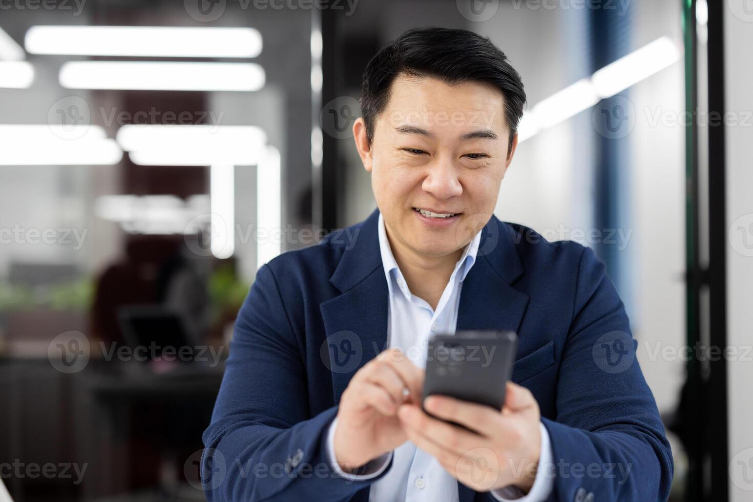 Asian man inside office with phone in hands, smiling contentedly, businessman using smartphone sitting at desk, mature boss in business suit browsing social media, typing text message call. photo