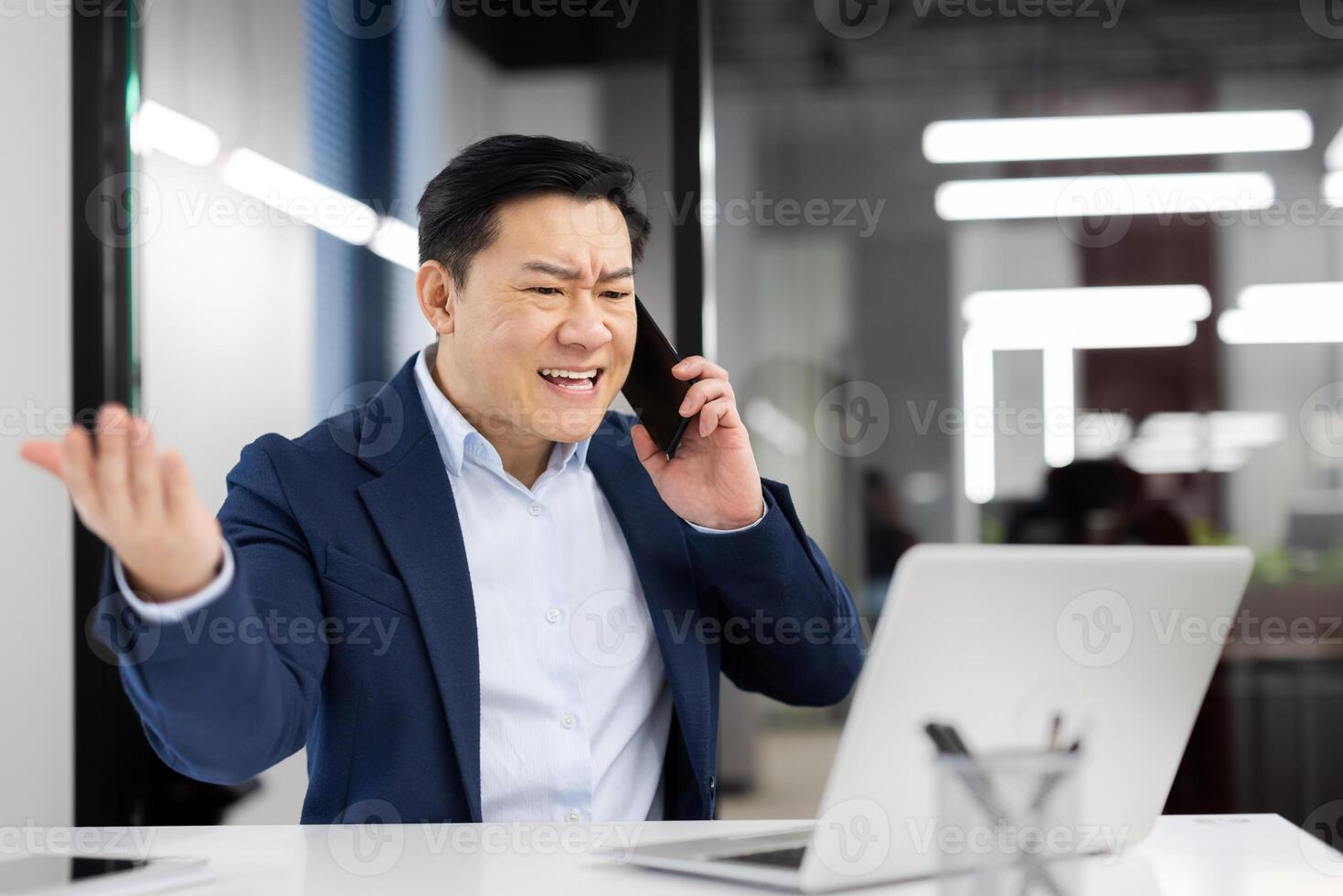 Angry grumpy boss shouting while talking on the phone, experienced asian man in business suit with phone in hands working inside office with laptop, annoyed businessman. photo