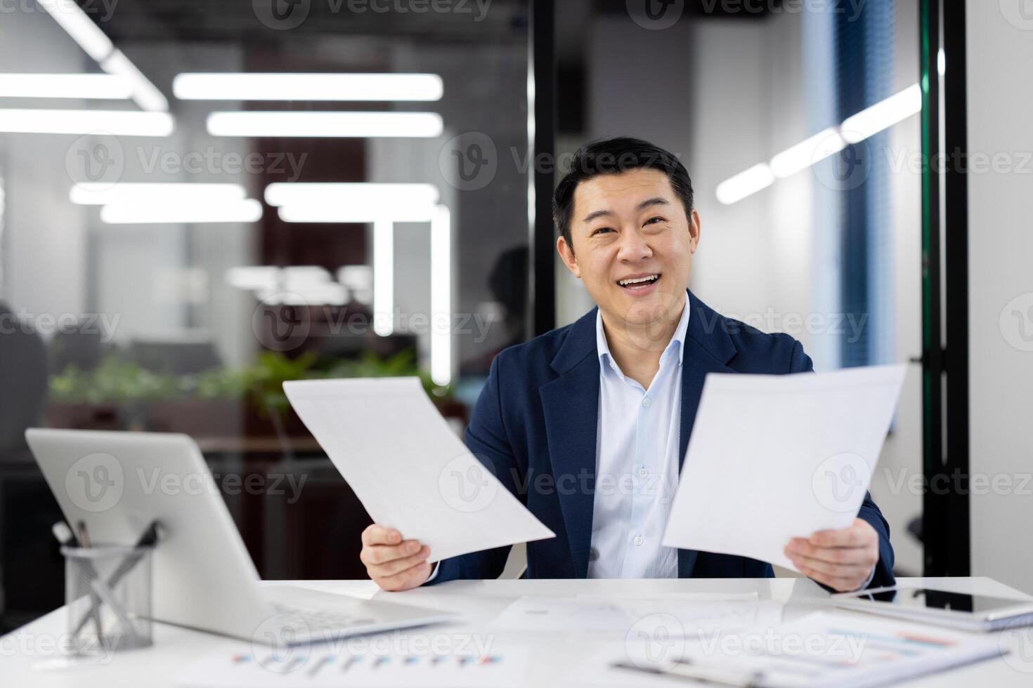 Happy Asian businessman holding papers with a pleased expression in a contemporary office setting, showing success and confidence. photo