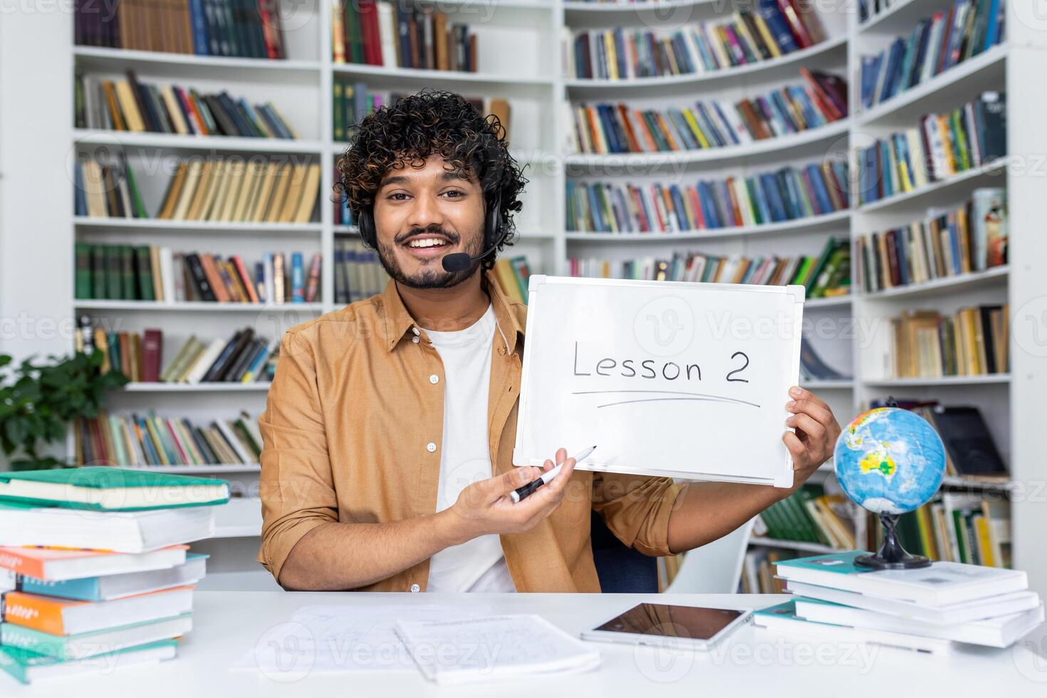 A young, engaging male teacher with a headset smiles as he presents 'Lesson 2' written on a whiteboard, surrounded by stacks of books in a bright library environment, symbolizing education photo