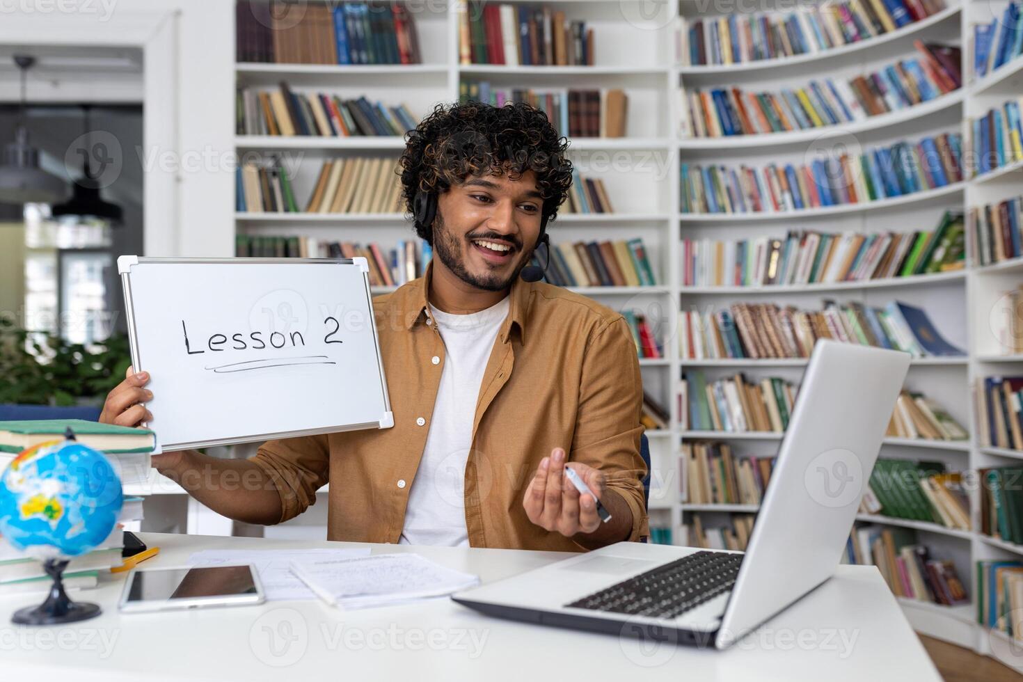 Latin American teacher teaches online students, the man sits in the library of the academic university with books, shows the subject of the lesson to the laptop camera. photo