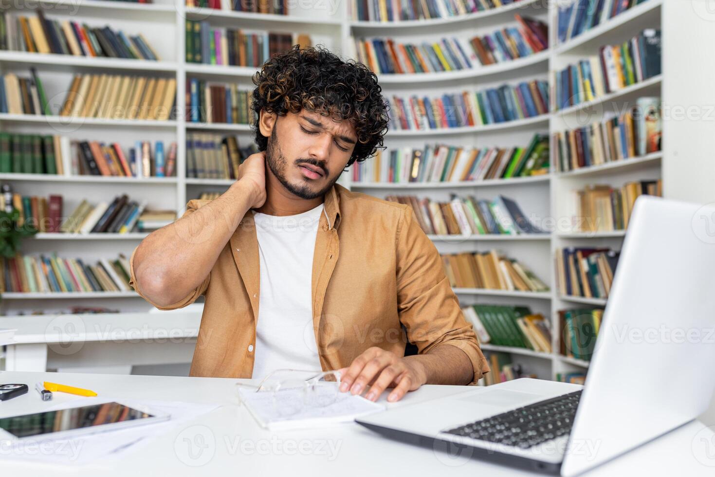 Suffering middle eastern male touching neck with hand while sitting by desktop with gadgets on background of bookshelves. Exhausted man struggling with results of seat with incorrect posture. photo