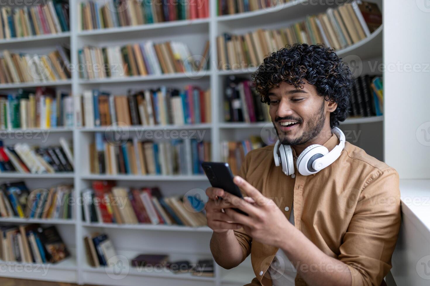 Young hispanic student smiling and happy, using online learning app, smiling man in headphones sitting inside academic library among books. photo
