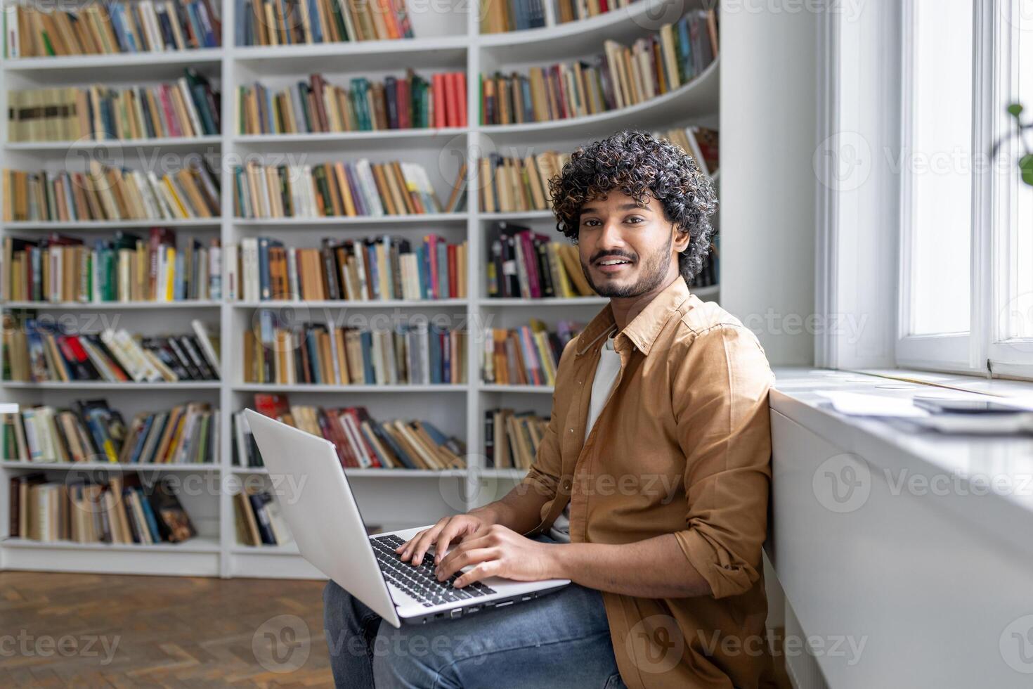 An engaged young adult sits comfortably in a well-lit space, surrounded by books, focused on work being done on a laptop. Setting suggests a modern library or a study room with a peaceful atmosphere. photo