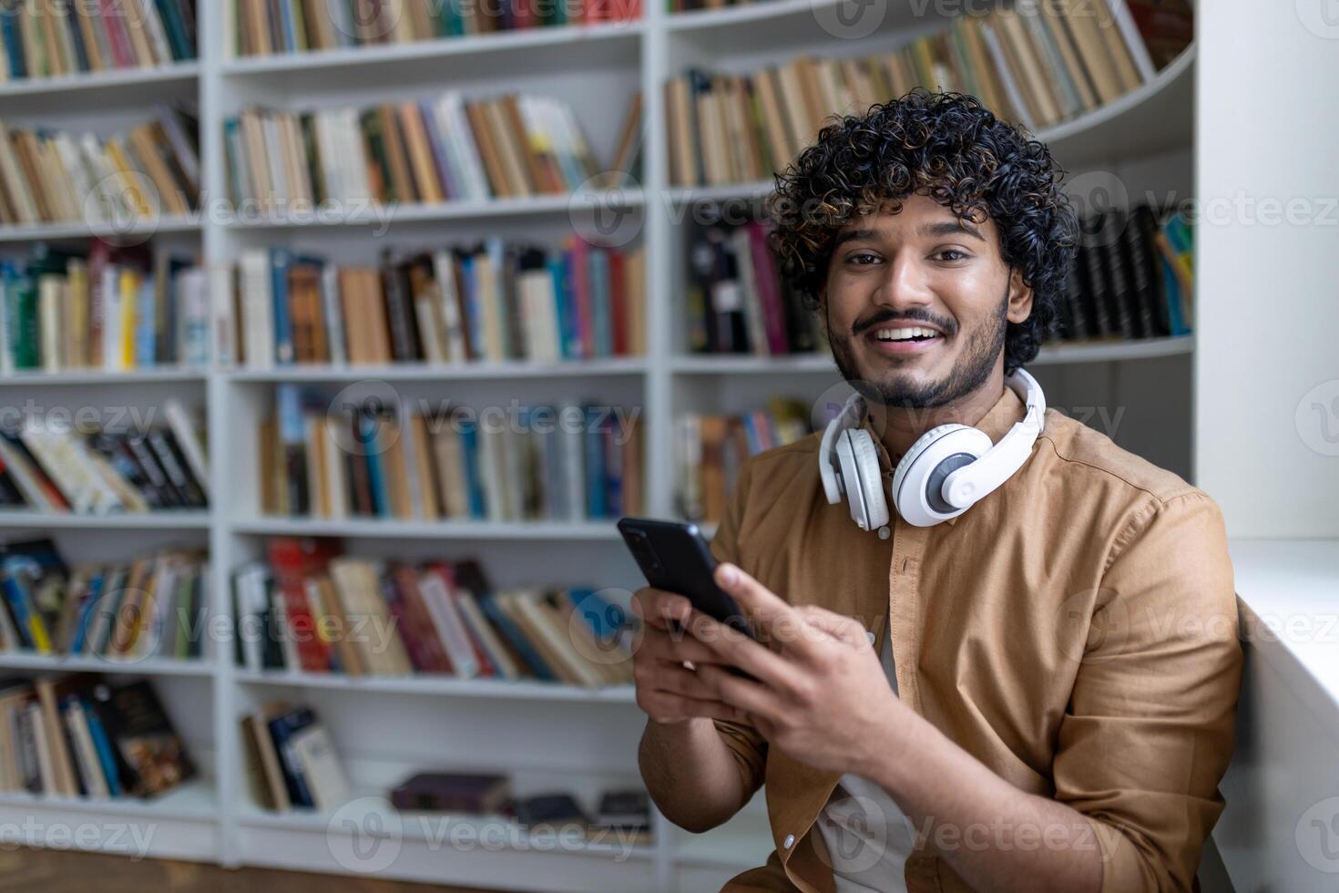 Young hispanic student inside public library studying, smiling and looking at camera, man holding phone, using study app, with headphones among shelves. photo