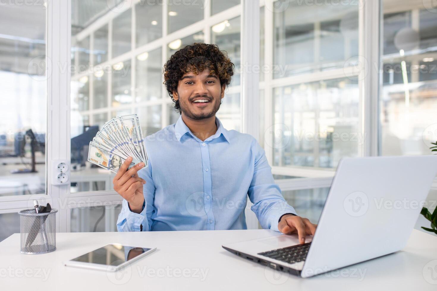 Successful financial analyst waving fan of dollar bills and looking at camera by desktop with portable computer. Young businessman enjoying results of productive work and trading on stock exchange. photo
