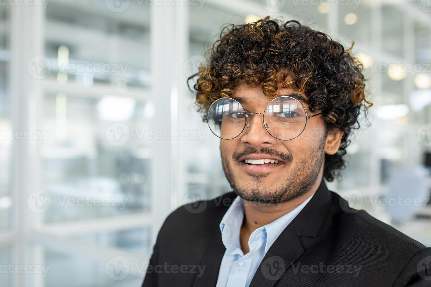 A young man with curly hair and glasses smiles confidently while dressed in a smart suit, positioned in a bright, contemporary office environment. photo