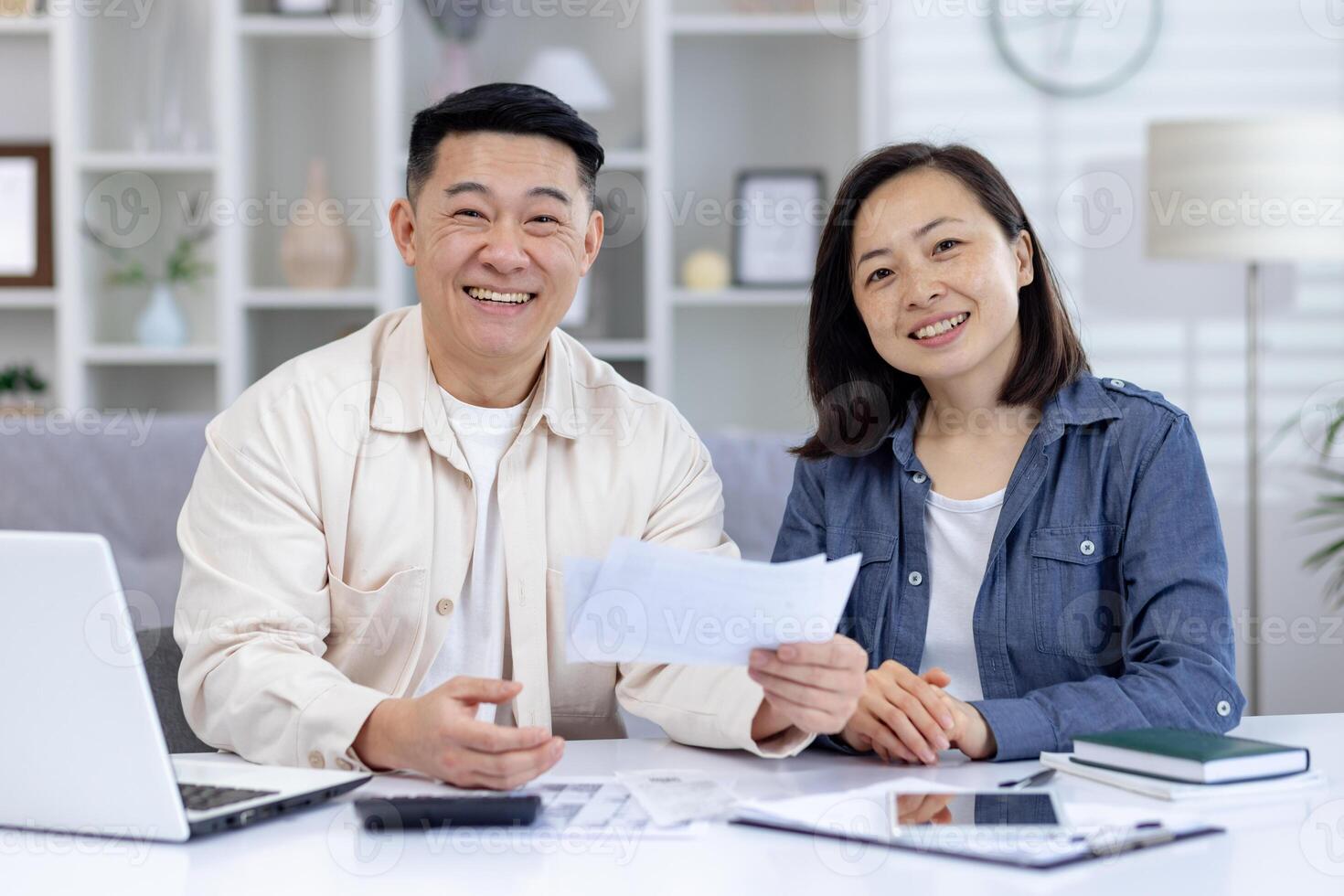 Happy asian couple sitting at table managing their finances using a laptop and documents, showing a positive family bond. photo