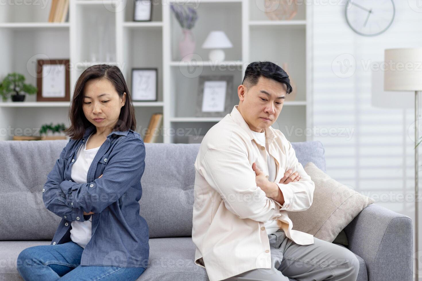 Asian couple feeling upset and distant while sitting on a couch in a living room. Emotions of conflict and disagreement are apparent between them. photo