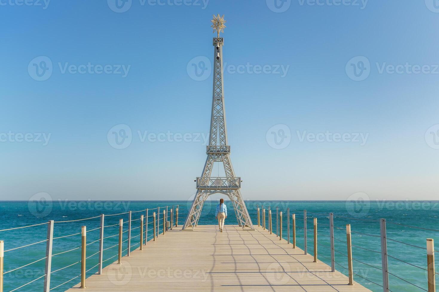 Large model of the Eiffel Tower on the beach. A woman walks along the pier towards the tower, wearing a blue jacket and white jeans. photo